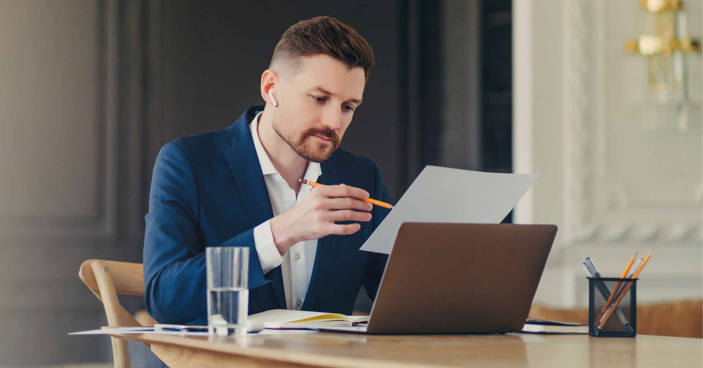 Business professional reviewing a document while working on a laptop at a desk.